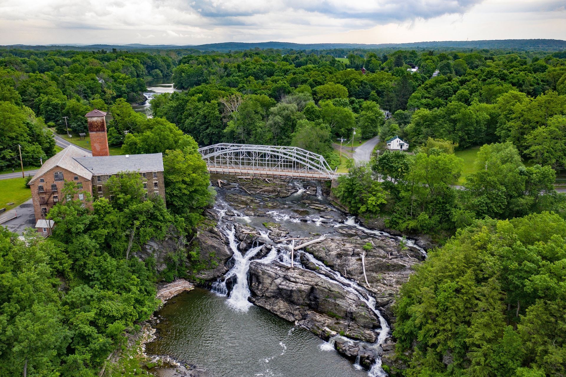 Aerial of a river and waterfall with a steel bridge and brick building. Mountains can be seen in the background