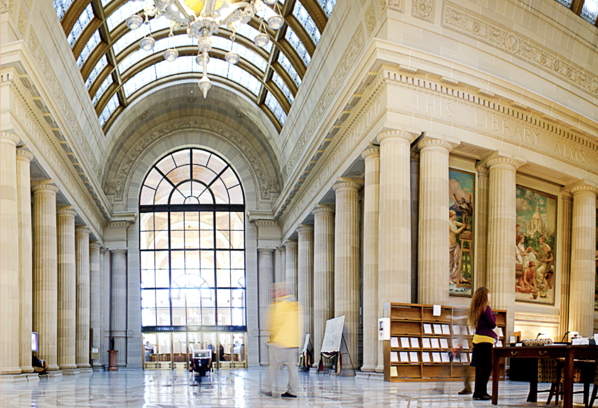 Inside a room with tall ceilings and lined by columns. The room has a glass dome ceiling and a large window.